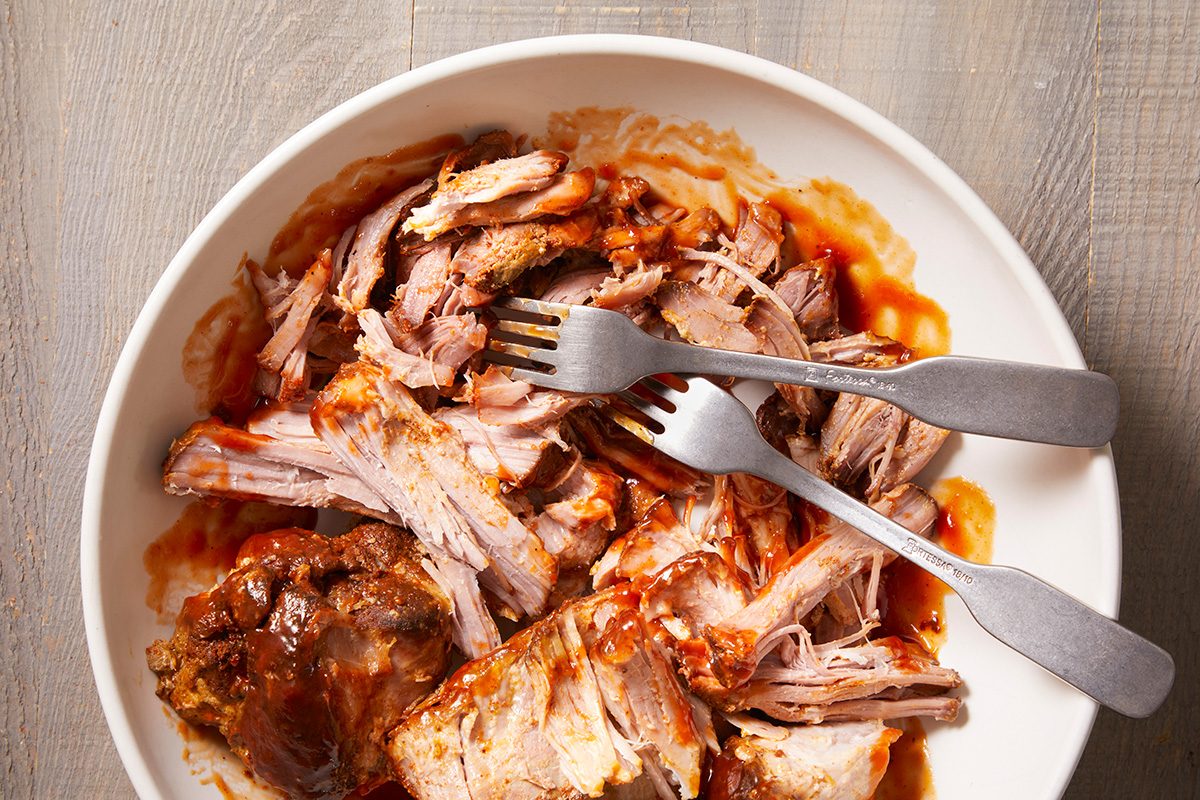 Shredded pieces of cooked pork in a white bowl, with two forks pulling apart the meat; some barbecue sauce is visible on the meat and bowl. The bowl is on a light wooden surface.