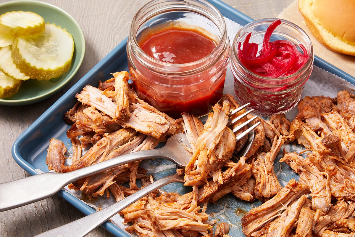 A tray with shredded pulled pork, two forks, a jar of barbecue sauce, a jar of pickled red onions, and a side plate with sliced pickles.