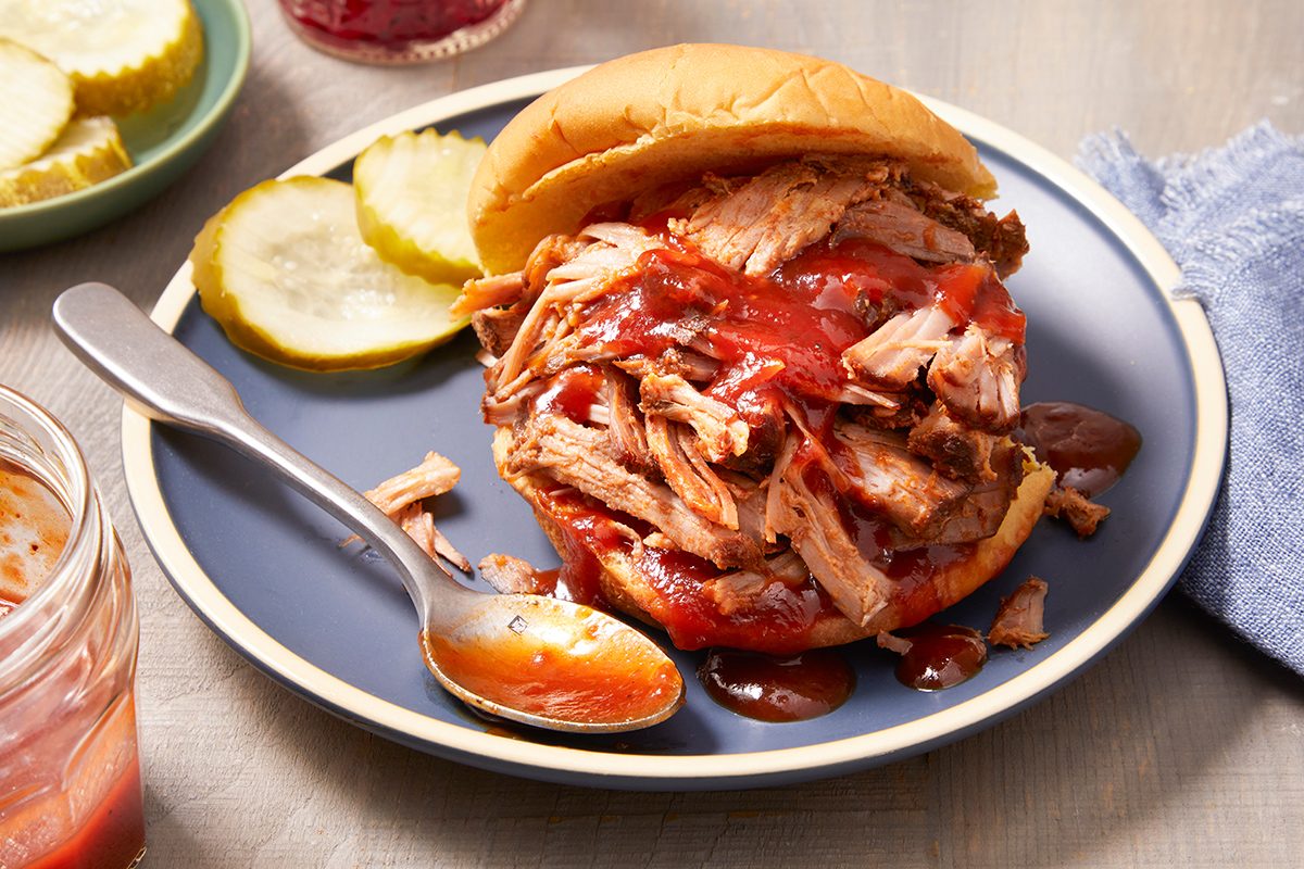 A pulled pork sandwich with barbecue sauce on a bun sits on a blue plate, accompanied by pickle slices and a spoon with sauce. A jar of sauce and a plate of pickles are nearby.