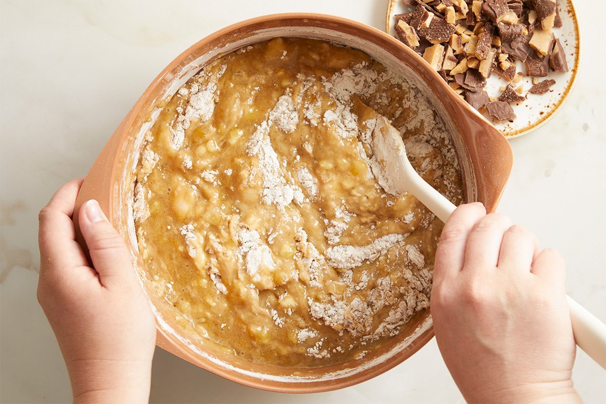 Overhead horizontal image of Elise Jesse's Bourbon Banana Bread. Hands mix the batter in a bowl with a wooden spoon, flour coats the top of the batter. Crushed Heath candy bars sit on a plate to the side.