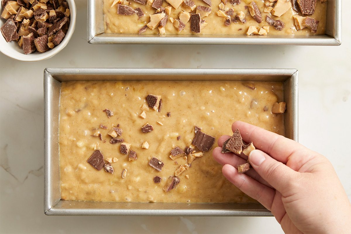 Overhead horizontal image of batter in two metal loaf pans being topped with crushed Heath candy bars. A hand is shown adding the candy bars to the batter, while a separate bowl of Heath candy bar pieces sits nearby.