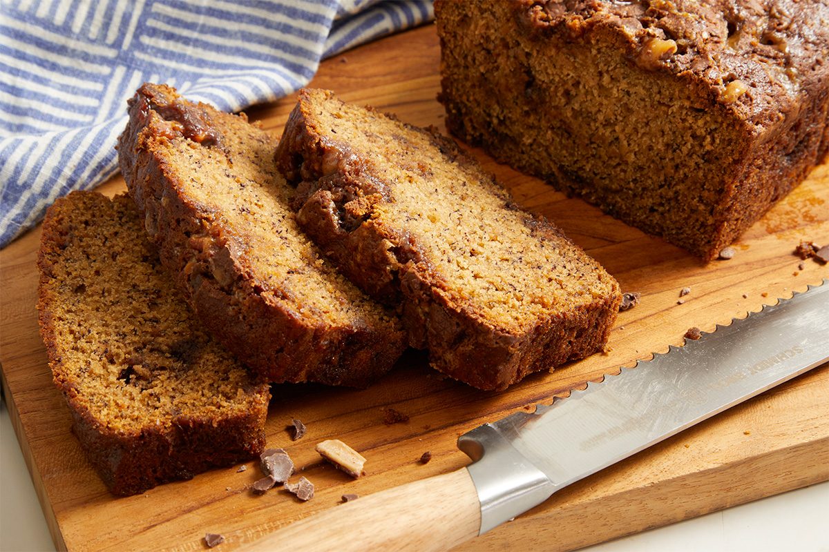 Eye Level horizontal image of sliced Bourbon Banana Bread on a wooden cutting board with a bread knife. The loaf is sliced and the inside texture is visible with bits of crushed Heath candy bars. A blue striped dish towel is slightly visible in the background.