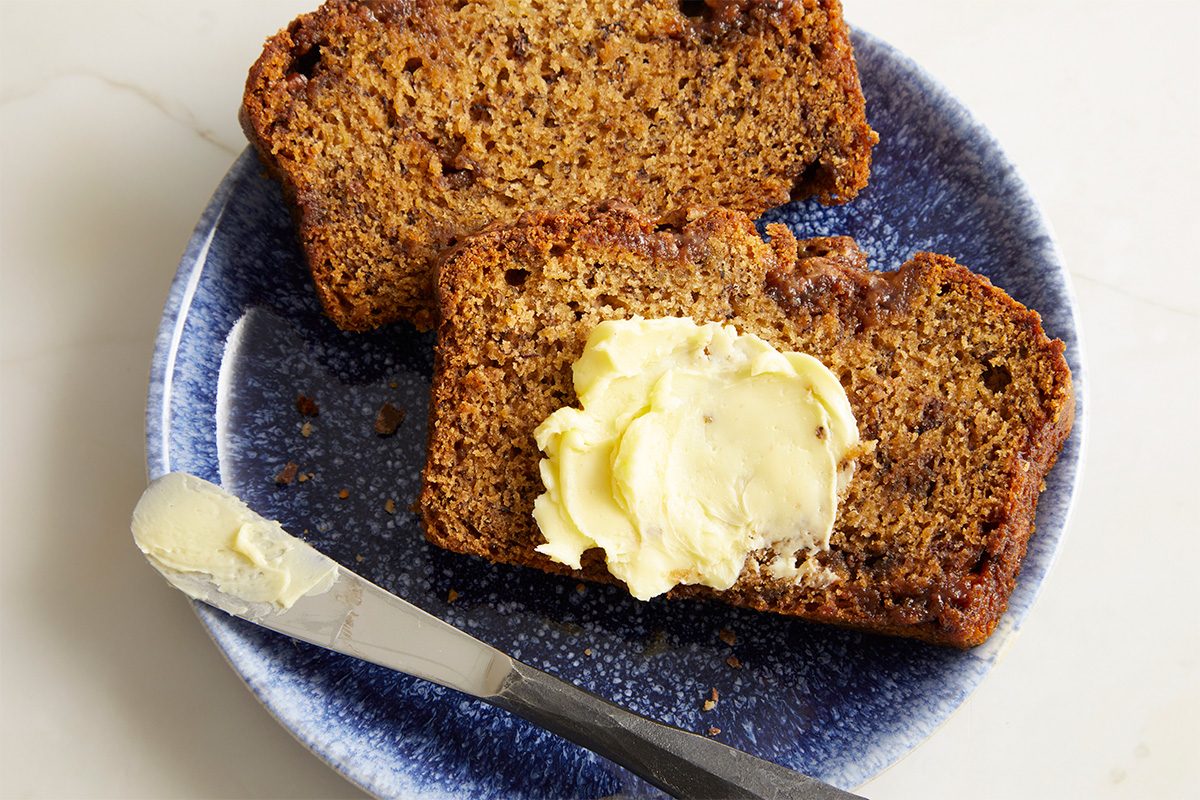 Overhead horizontal image of sliced banana bread with butter on a blue plate, with additional butter on a knife. Two slices of Elise Jesse's Bourbon Banana Bread are shown; one is topped with butter, and one slice is partially visible in the upper left corner. The butter knife has a portion of butter on it.