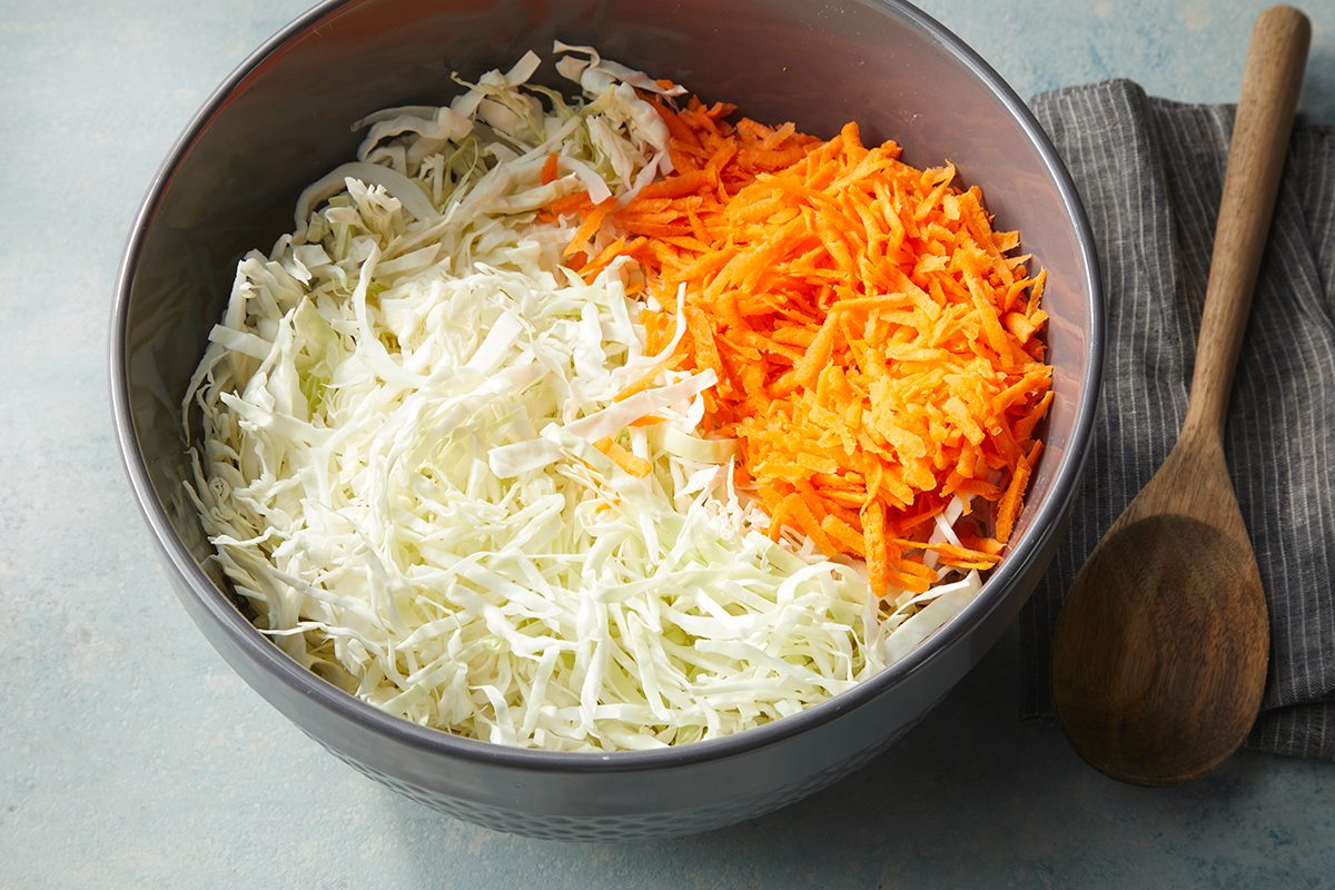 A metal bowl containing shredded cabbage and shredded carrots, placed on a gray surface next to a wooden spoon and a striped cloth napkin.