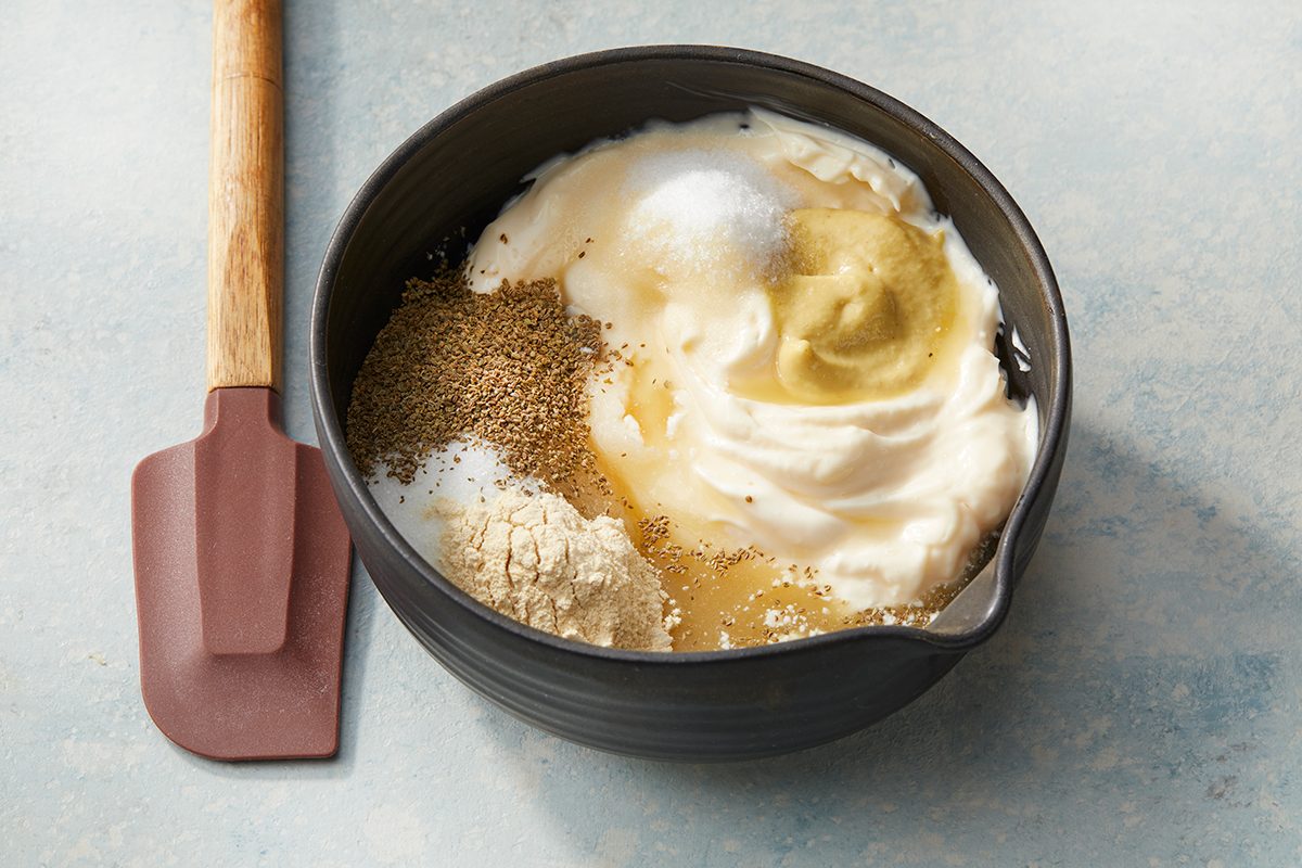 A black mixing bowl filled with yogurt, mustard, spices, and seasonings sits on a light surface next to a brown silicone spatula with a wooden handle.