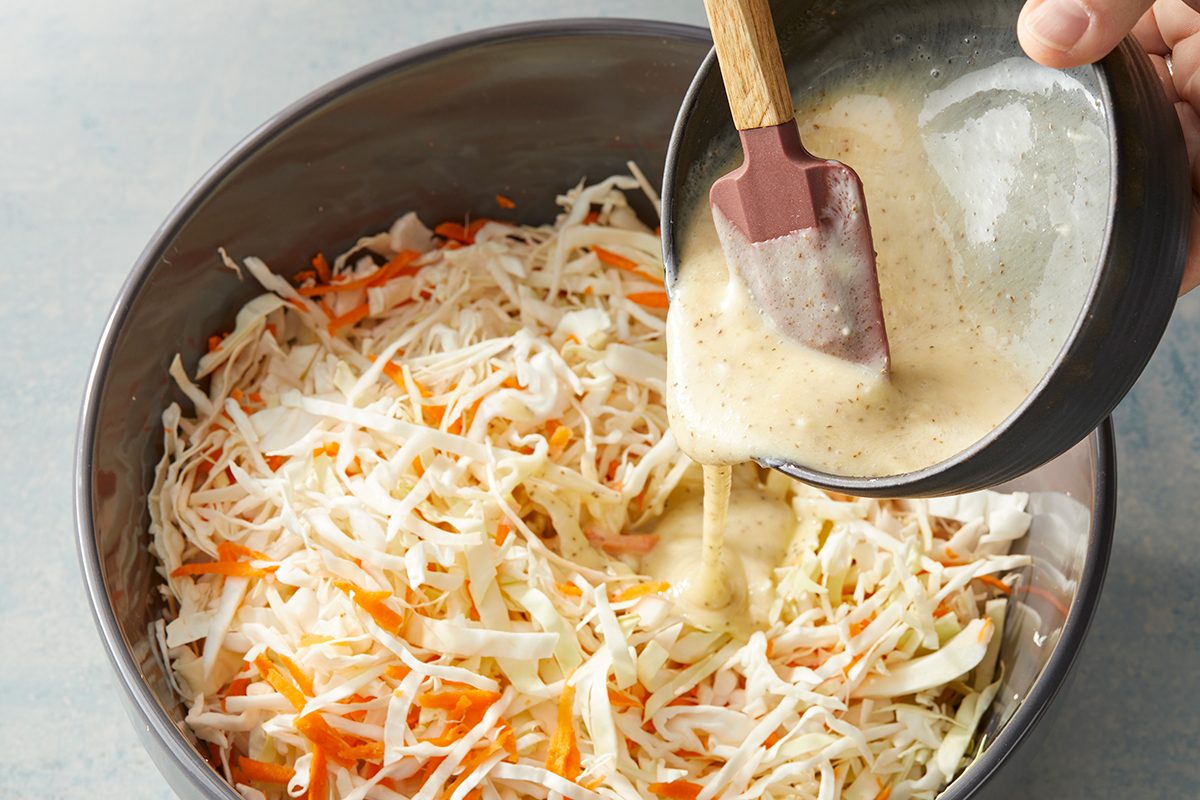 A bowl of shredded cabbage and carrots is being mixed as creamy dressing is poured over the vegetables with a spatula.