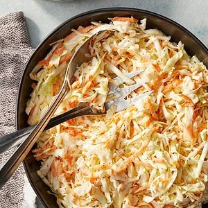 A bowl of coleslaw made with shredded cabbage and carrots, with metal tongs resting on the side. The bowl sits on a light surface next to a textured napkin.