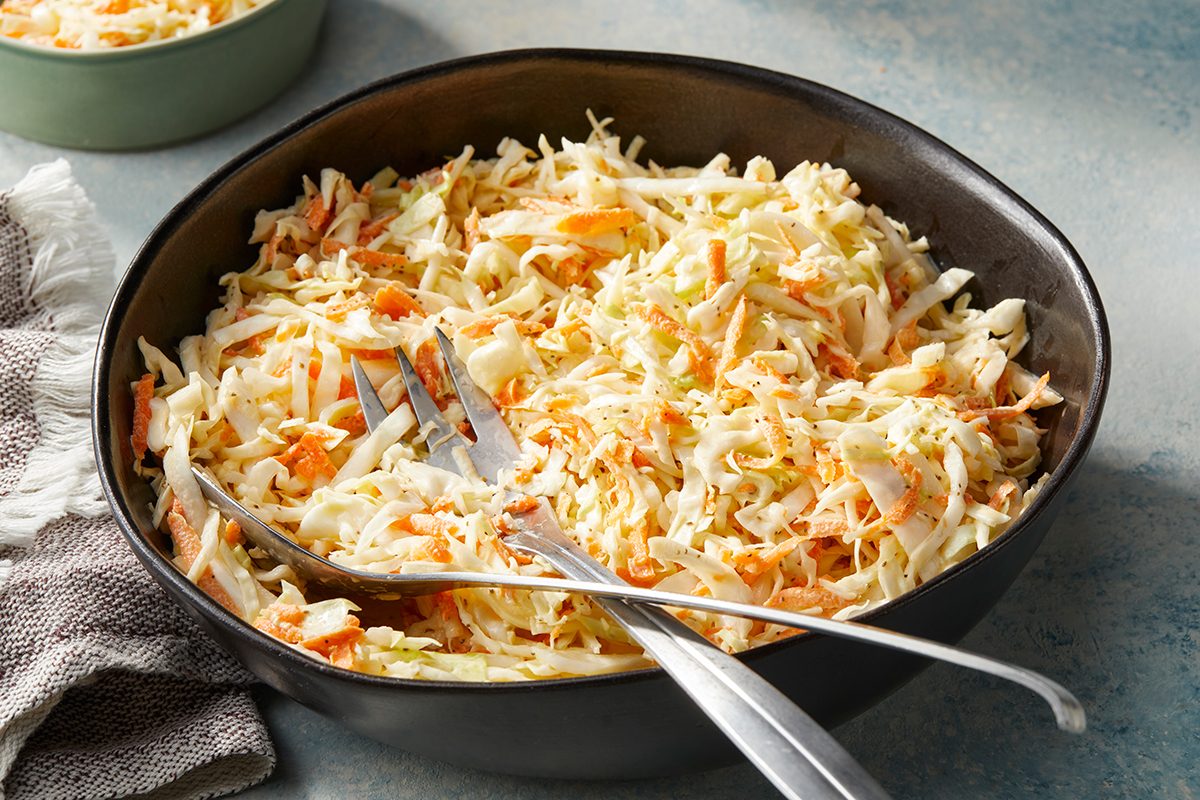 A black bowl filled with creamy coleslaw made of shredded cabbage and carrots, with serving utensils resting inside. A napkin is placed beside the bowl on a light-colored surface.