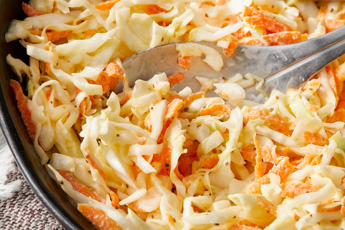 A close-up of a bowl of coleslaw, featuring shredded cabbage and carrots, with metal tongs resting in the mixture.