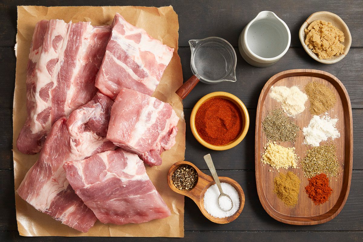Overhead horizontal image of raw pork baby back ribs, smoked paprika, brown sugar, salt, coarsely ground pepper, garlic powder, onion powder, ground cumin, ground mustard, dried thyme, dried oregano, cayenne pepper, white vinegar, and water.