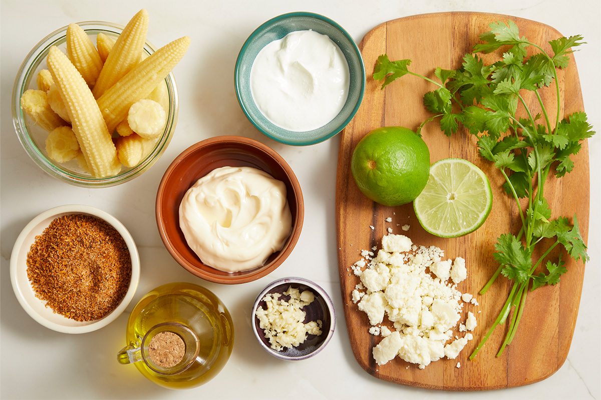 Overhead horizontal image of various ingredients laid out for a recipe on a white marble surface and a wooden cutting board. Bowls of baby corn, sour cream, mayonnaise, minced garlic, and a spice blend are visible alongside a bottle of olive oil. A whole lime, a lime half, crumbled cheese, and fresh cilantro are arranged on the cutting board.