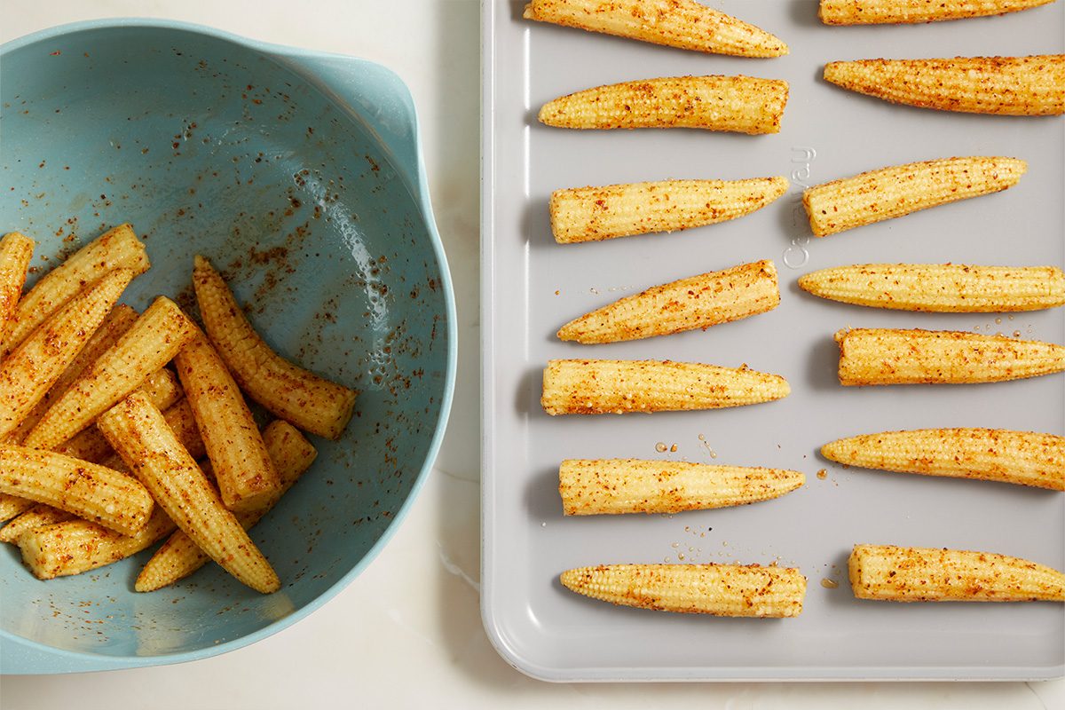 Overhead horizontal image of seasoned baby corn. On the left, a light blue mixing bowl holds baby corn coated in spices. On the right, multiple pieces of seasoned baby corn are arranged in rows on a grey baking sheet, ready for cooking.
