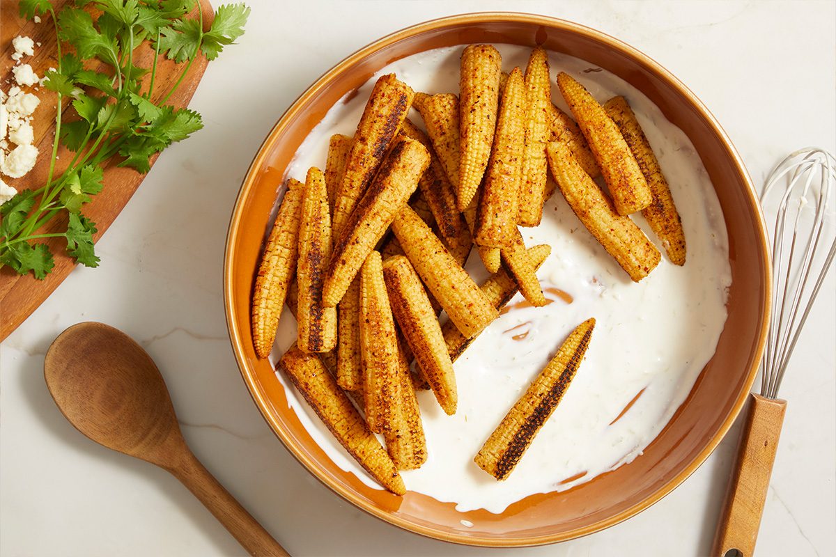Overhead horizontal image of seasoned baby corn arranged in a creamy white sauce within a brown ceramic bowl. On the left, a wooden cutting board features crumbled white cheese and fresh cilantro, with a wooden spoon and whisk also visible on the white marble surface.