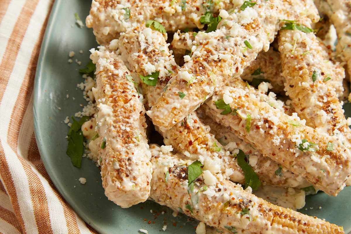 Overhead horizontal image of multiple short segments of corn on the cob piled on a light blue-green plate. Each piece is coated in a creamy white sauce, generously sprinkled with crumbled white cheese, fresh green cilantro, and red chili flakes.