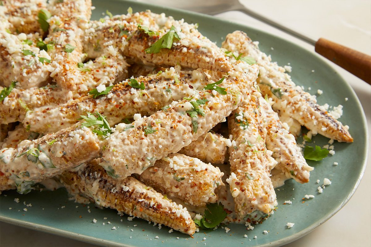 Eye Level horizontal image of a pile of mini corn cobs generously coated in a creamy white sauce. The corn is sprinkled with crumbled white cheese, chopped fresh cilantro, and red chili flakes, presented on a light teal plate.