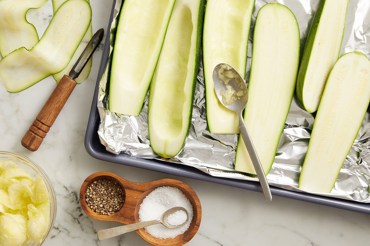 Zucchini halves with centers scooped out sit on a foil-lined baking sheet. Nearby are a spoon, a peeler, a bowl of zucchini flesh, and a small wooden dish with salt and pepper on a marble countertop.