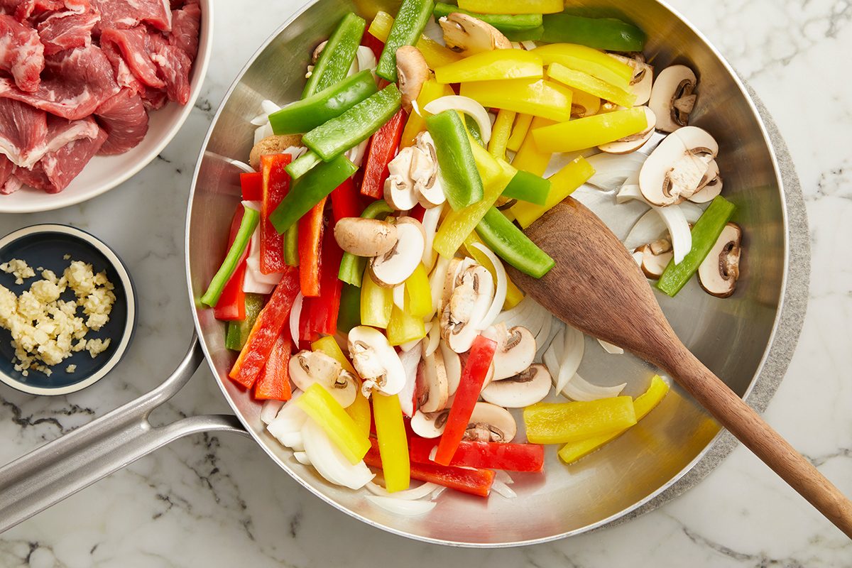 A skillet with sliced red, yellow, and green bell peppers, mushrooms, and onions being sautéed with a wooden spoon. Nearby are bowls of raw beef strips and minced garlic on a marble countertop.