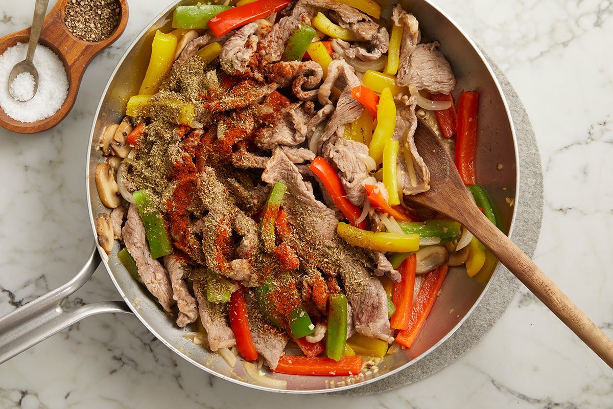 A skillet on a marble countertop filled with sliced beef, red, yellow, and green bell peppers, mushrooms, and seasoning. A wooden spoon rests in the pan; salt and pepper containers sit nearby.