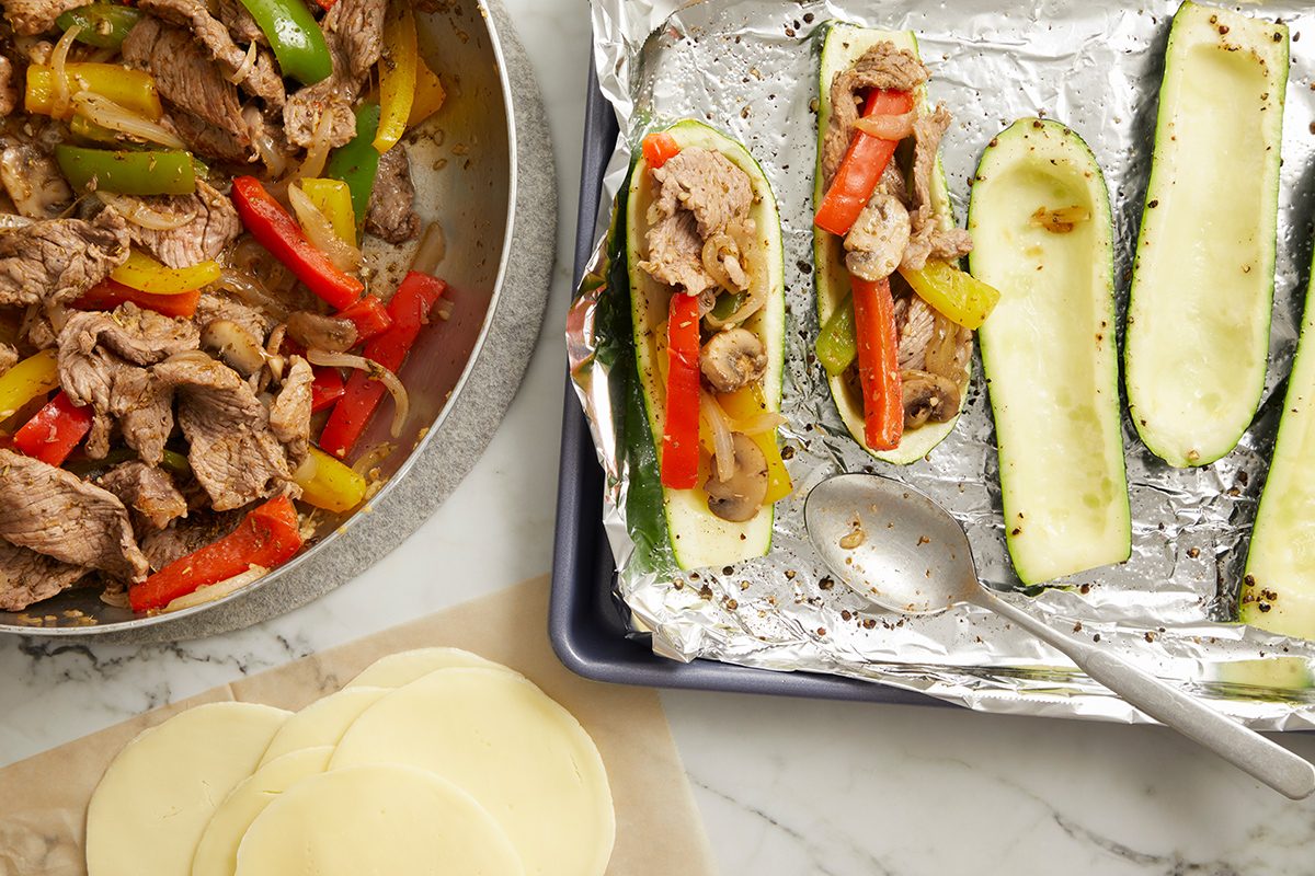 Sliced cooked beef and colorful bell peppers in a pan beside zucchini boats being filled with the mixture on a foil-covered tray; slices of cheese are on the side.