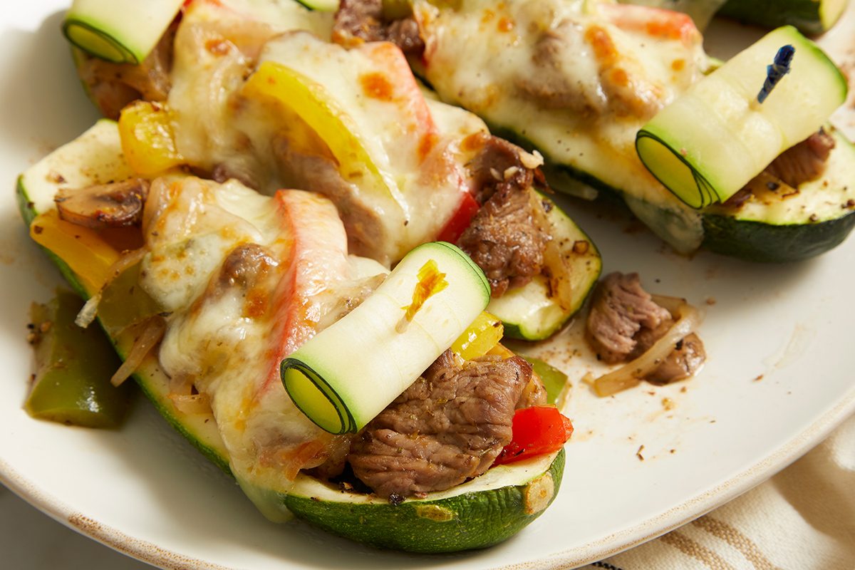 Stuffed zucchini boats filled with cooked beef, bell peppers, and onions, topped with melted cheese and garnished with thin zucchini ribbons, served on a white plate.