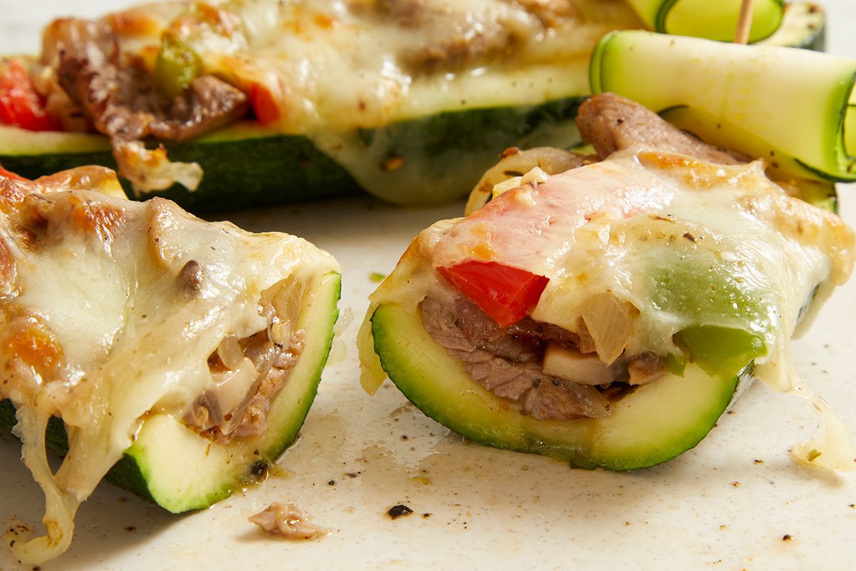 Close-up of zucchini boats stuffed with sliced beef, bell peppers, and onions, topped with melted cheese. One zucchini boat is cut open, showing the filling. Food is on a light-colored surface.