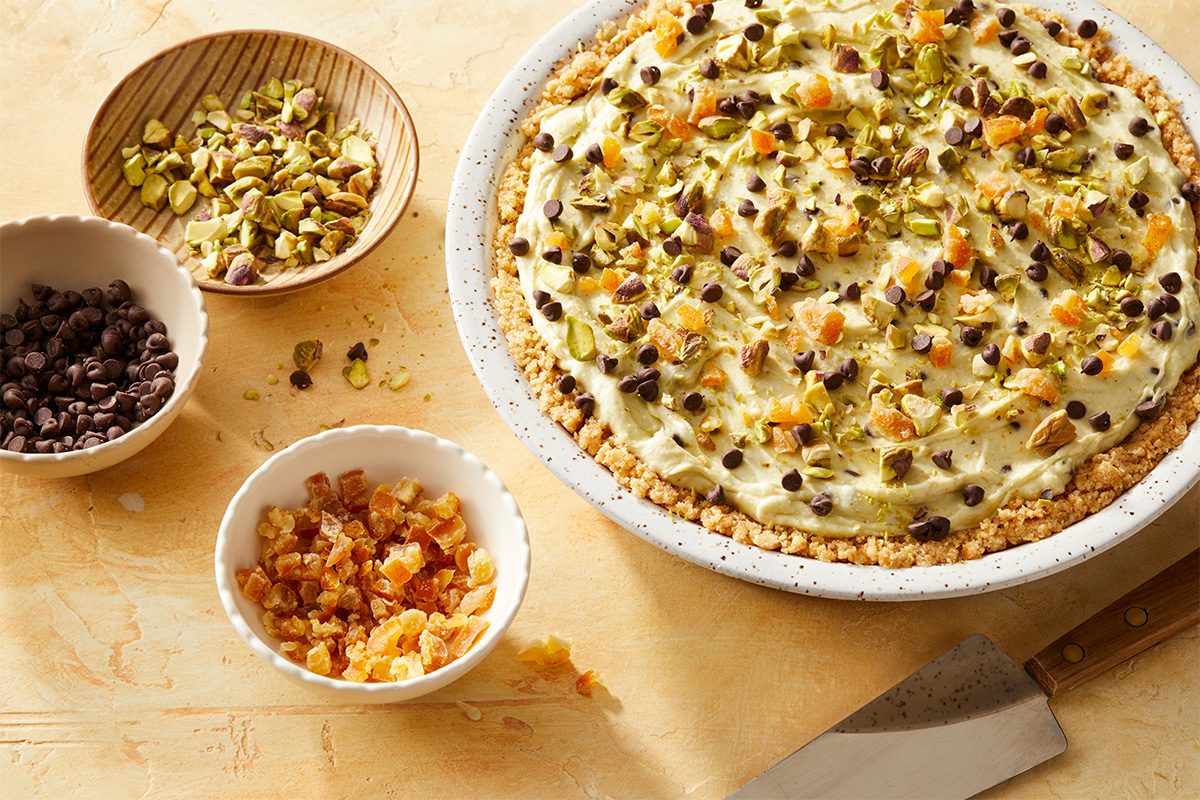 Overhead horizontal image of a dessert pie in a white ceramic dish, generously topped with chopped pistachios, mini chocolate chips, and diced dried fruit. Three small bowls containing additional pistachios, chocolate chips, and dried fruit are arranged next to the pie, with a pie server visible at the bottom right. | [Original] RMS 282849 Pistachio Cannoli Pie; How to; Step 3; have toppings propped and start of adding chopped pistachios