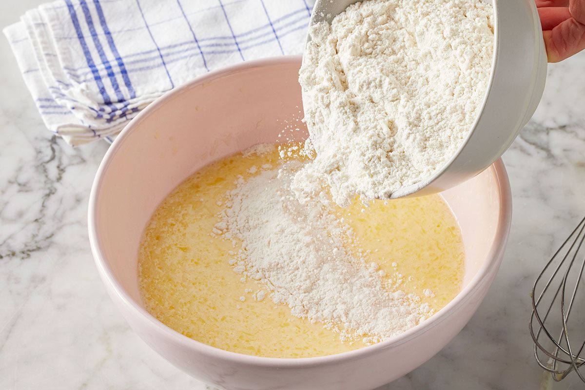 Eye level horizontal image of a hand pouring white flour from a small white bowl into a pink mixing bowl filled with a yellow liquid mixture. A whisk with a wooden handle and a white and blue checkered towel are also visible on the marble countertop.