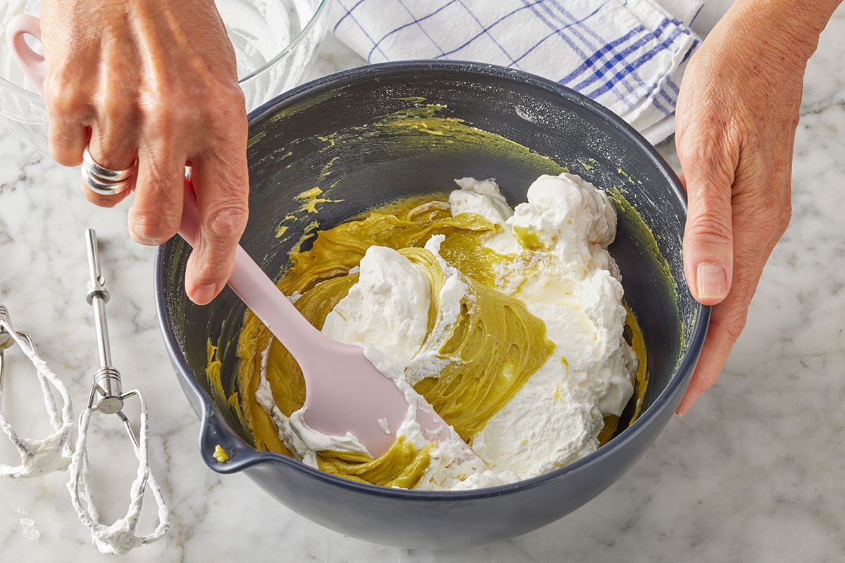 Overhead horizontal image of a person's hands using a pink spatula to gently fold fluffy white whipped cream into a pale green creamy mixture within a dark gray mixing bowl. In the background, mixer beaters rest on a white marble countertop next to a blue and white checkered towel.