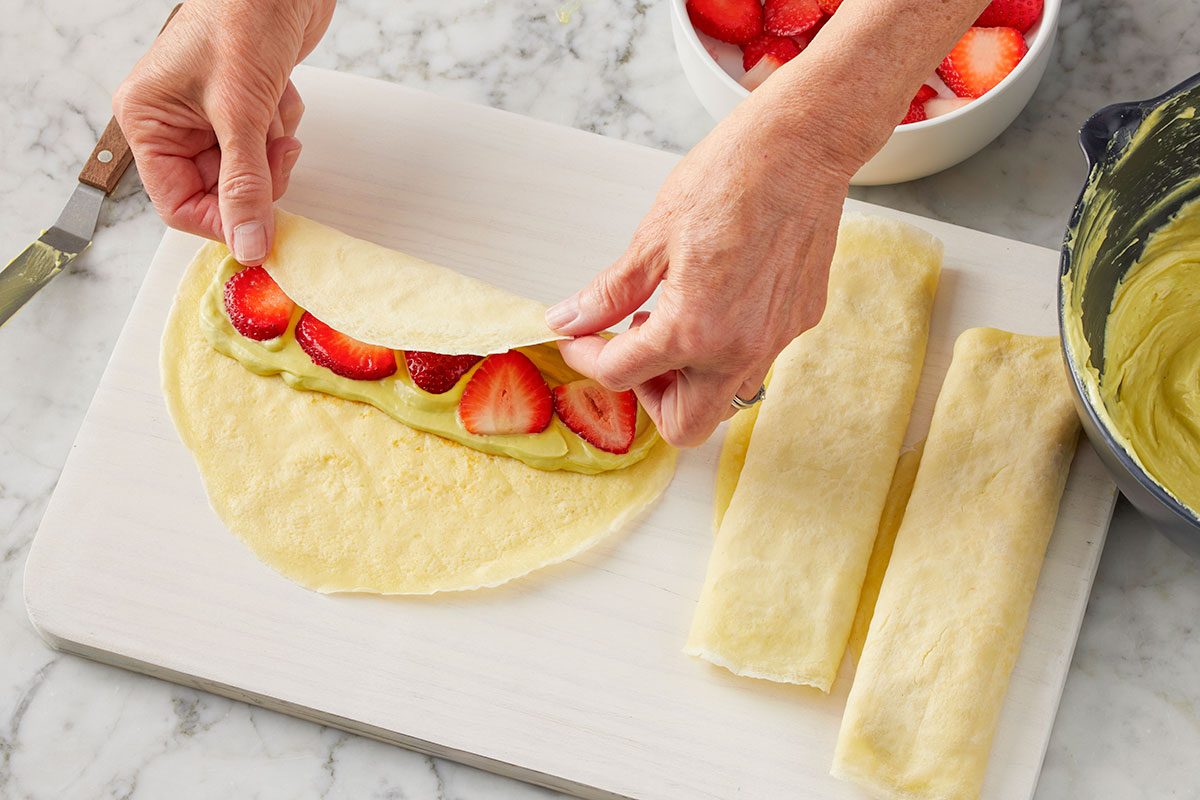 Overhead horizontal image of hands rolling a crepe filled with green avocado cream and sliced strawberries on a white cutting board. Two finished rolled crepes are beside it, with a bowl of additional sliced strawberries and a bowl of the green cream in the background.