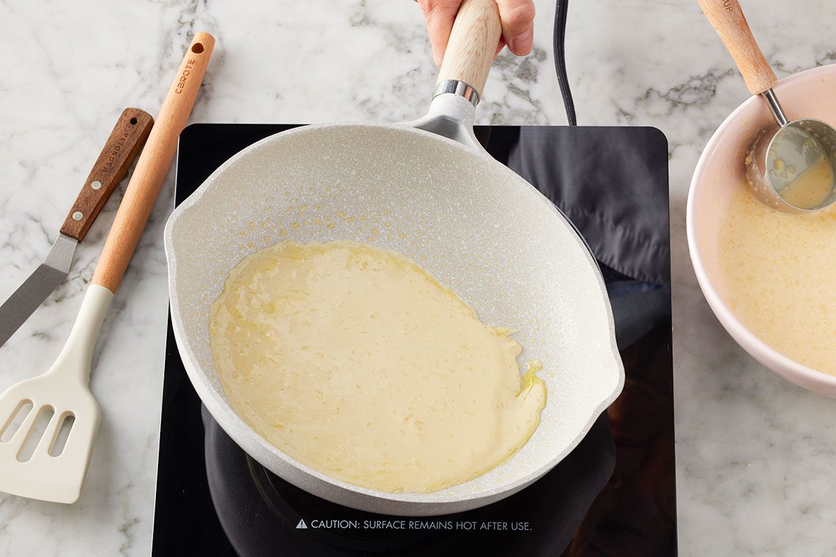 Overhead horizontal image of a hand holding a frying pan with a thin layer of light yellow batter cooking on an induction cooktop. A bowl of additional batter, a spatula, and an offset spatula are visible nearby on a marble surface.