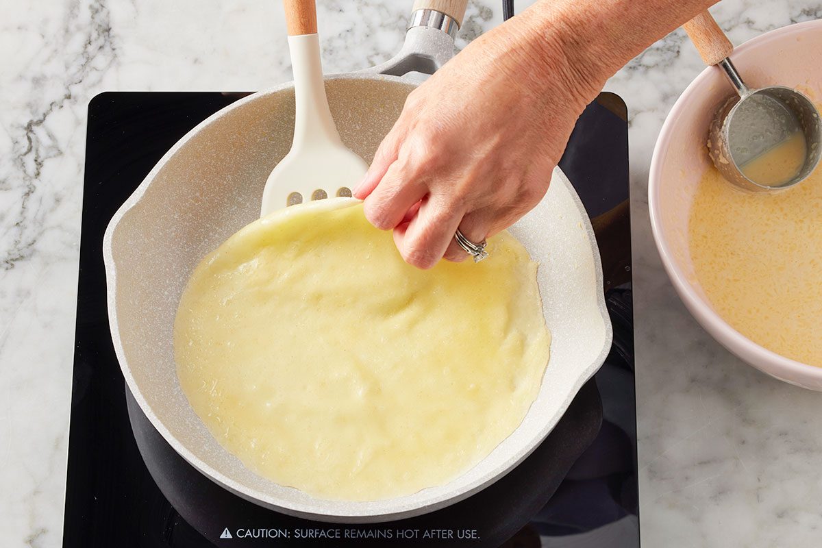 Overhead horizontal image of a hand using a white spatula to loosen the edge of a thin, light-yellow pancake or crepe cooking in a gray frying pan on a black stovetop. To the right, a pink bowl holds more pancake or crepe batter with a silver measuring cup dipped inside.