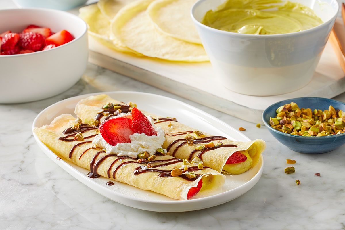 Eye Level horizontal image of two filled crepes presented on a white platter, topped with chocolate drizzle, whipped cream, sliced strawberries, and chopped pistachios. In the background, there are bowls of fresh chopped strawberries, plain crepes, a bowl of green pistachio cream, and more chopped pistachios.