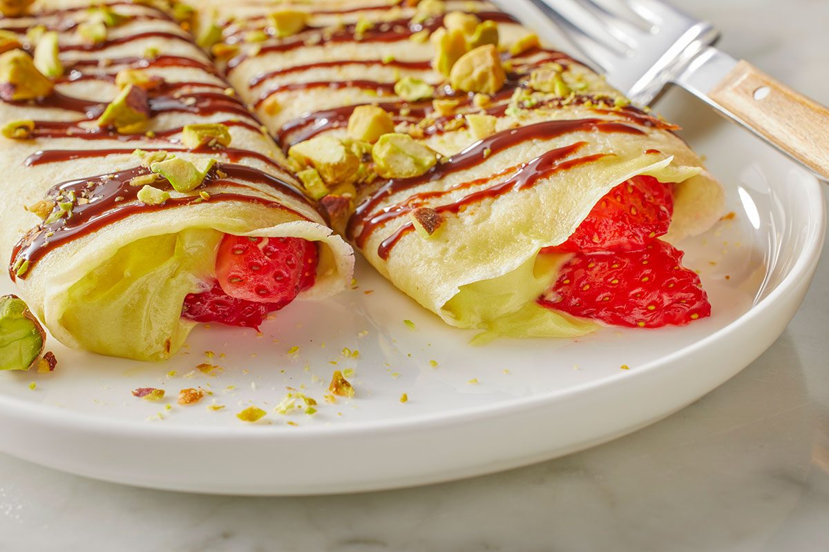 Eye Level horizontal image of two rolled crepes presented on a white plate, filled with creamy custard and fresh strawberries. The crepes are garnished with a drizzle of chocolate sauce and chopped pistachios.