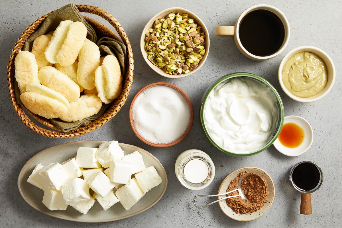 Overhead horizontal image of various dessert ingredients arranged on a gray surface. A basket of ladyfingers, bowls of chopped pistachios, granulated sugar, Greek yogurt, cream cheese cubes, cocoa powder, coffee, espresso, milk, pistachio cream, and vanilla extract are neatly displayed.