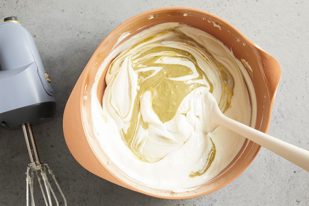 Overhead horizontal image of a light brown mixing bowl containing a white, creamy mixture being swirled with a green mixture. A light-colored silicone spatula is partially submerged in the mixture, and a light blue hand mixer with beaters is visible next to the bowl.