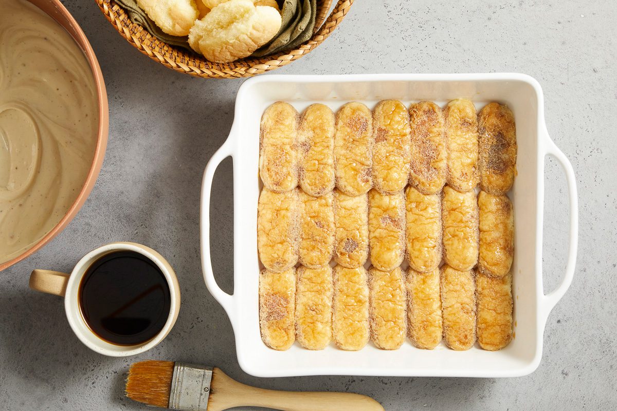 Overhead horizontal image of a white rectangular baking dish filled with ladyfingers, some coated with a sugary glaze. To the left, a bowl of light-colored cream and a mug of dark coffee are visible, along with a pastry brush. A basket holding more ladyfingers sits in the upper left corner.