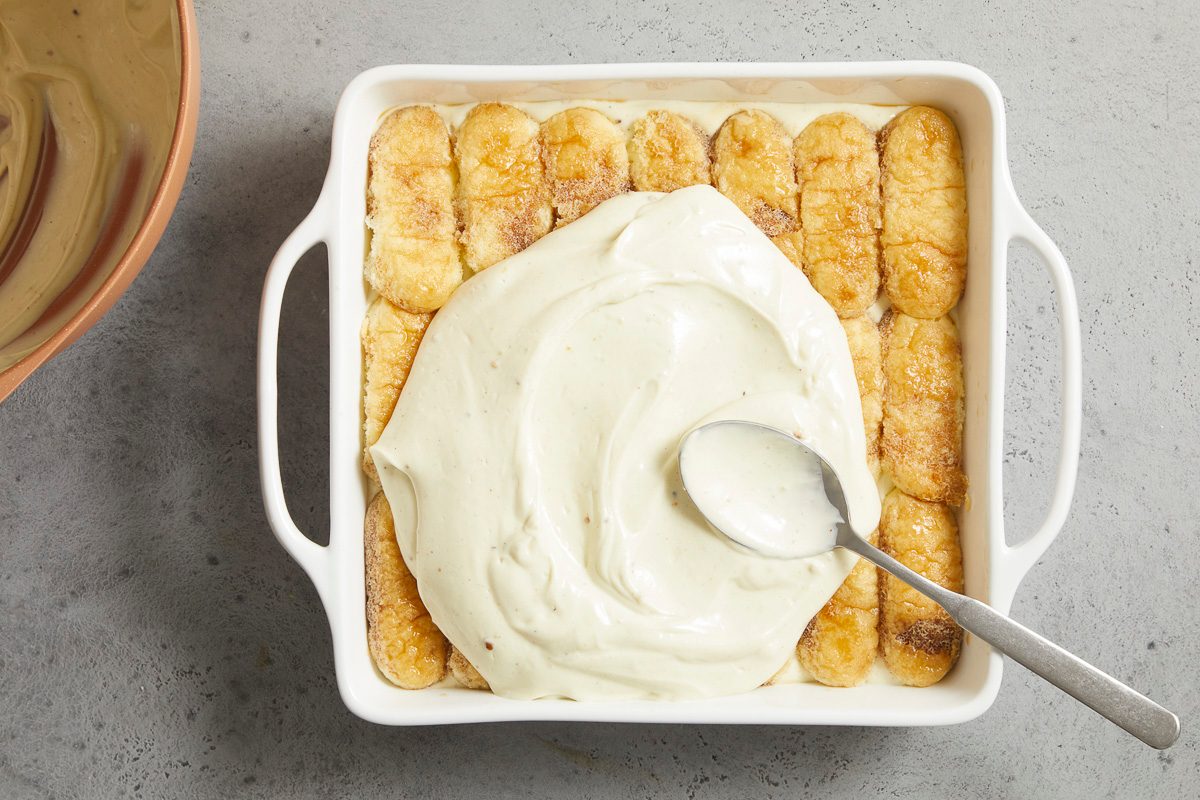 Overhead horizontal image of a white rectangular baking dish being assembled. Ladyfinger cookies, appearing soaked, line the bottom, with a spoon spreading a thick, light-colored creamy mixture over them.