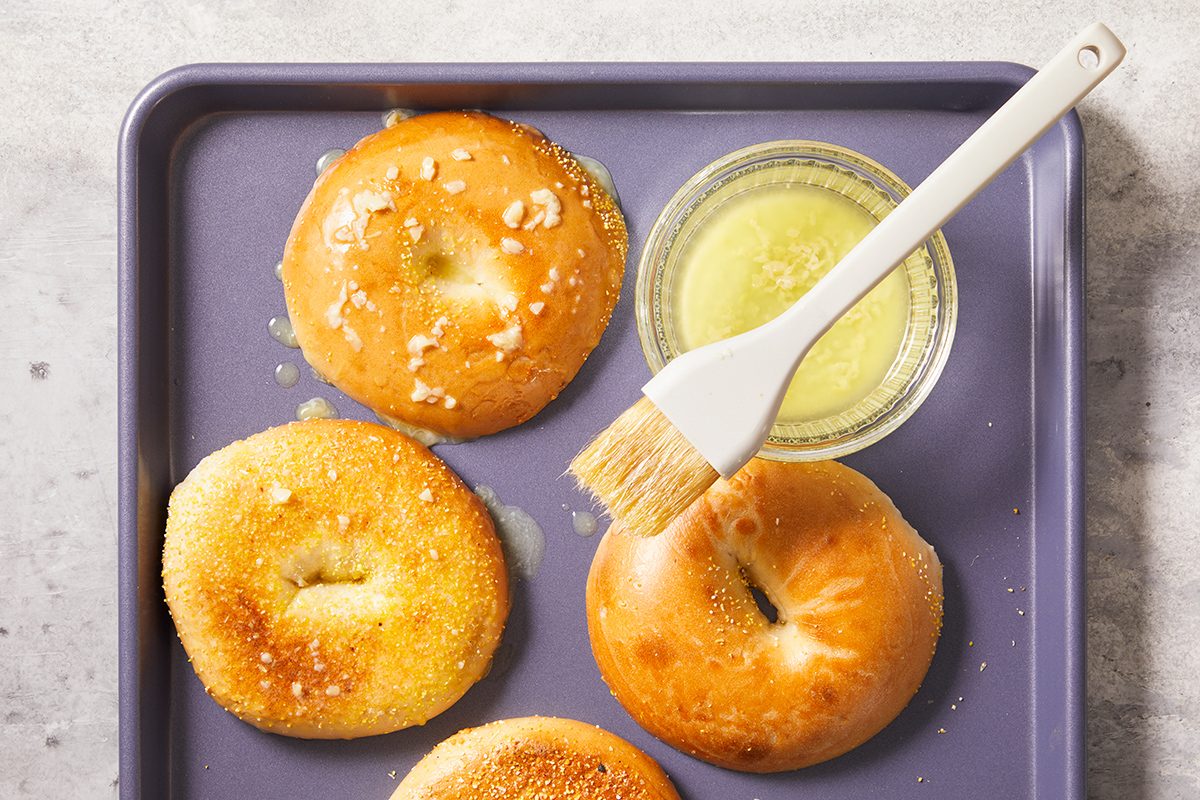 Four bagels on a baking tray, with a small bowl of melted butter and a pastry brush. Some bagels are shiny with butter, and others are sprinkled with coarse salt or seeds.
