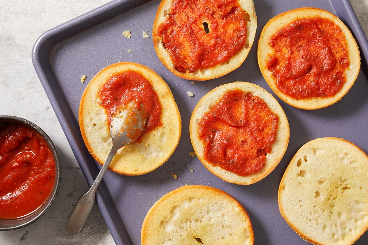 Six halved bagels on a baking tray, each spread with tomato sauce. A spoon rests on one bagel half, and an open can of tomato sauce sits nearby.