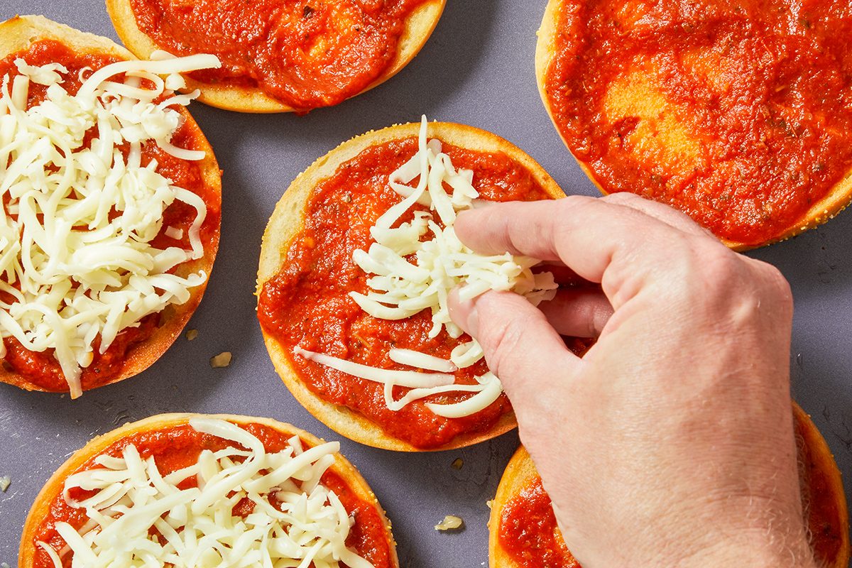 A hand sprinkles shredded cheese onto mini pizza crusts covered with tomato sauce, preparing homemade mini pizzas on a baking tray.