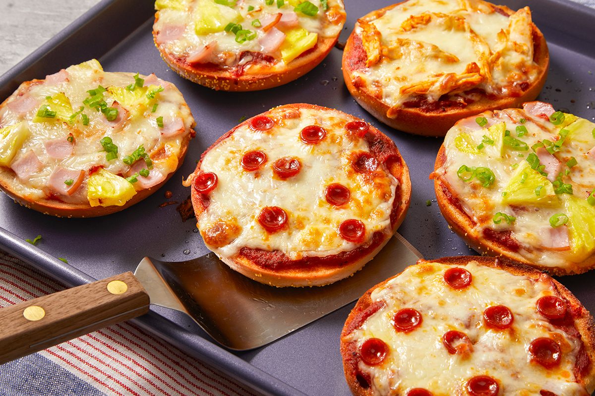 Six mini pizzas on a baking tray, three topped with melted cheese and mini pepperoni slices, and three with cheese, ham, pineapple, and green onions. A spatula is under one of the mini pepperoni pizzas.