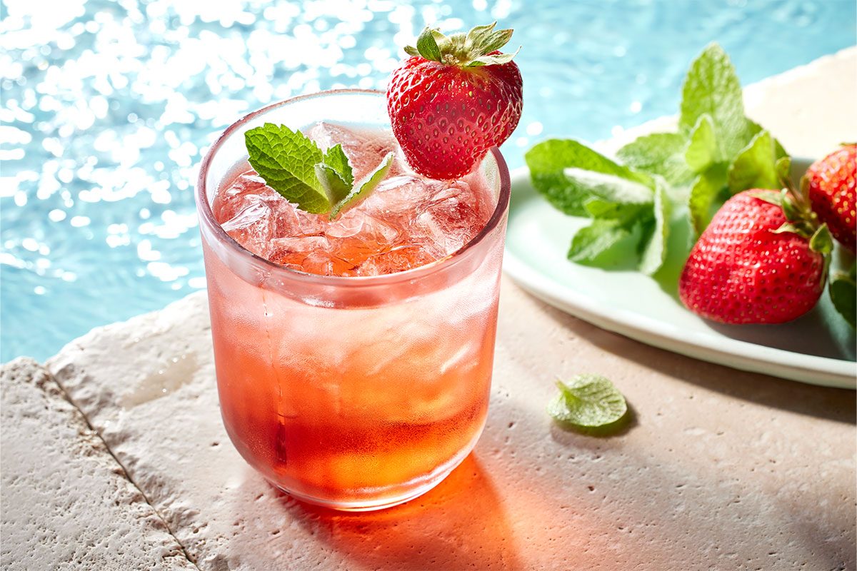 Eye Level horizontal image of a cold drink in a glass garnished with a strawberry and mint sprig. A plate of strawberries and mint sprigs sits in the background near the water.