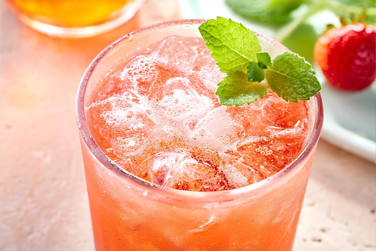 Eye Level horizontal image of a cocktail in a tall glass, garnished with mint. The cocktail contains ice and appears to have bubbles. A strawberry and another drink are visible in the background.