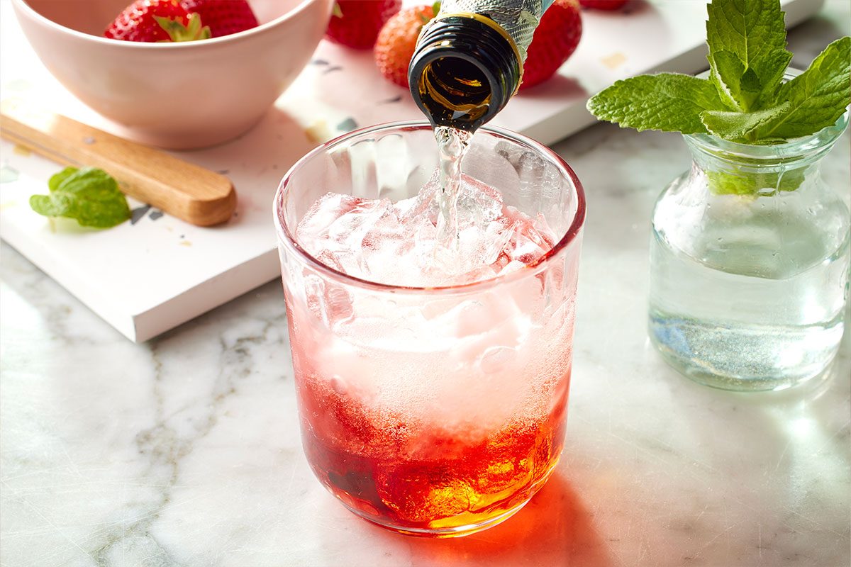 Eye Level horizontal image of sparkling wine being poured into a glass of ice and red-colored liquid. Strawberries are in the background, along with a sprig of mint in a small vase. | [Original] Fill large wine or rocks glass with ice. Add Prosecco, strawberry liqueur and club soda.