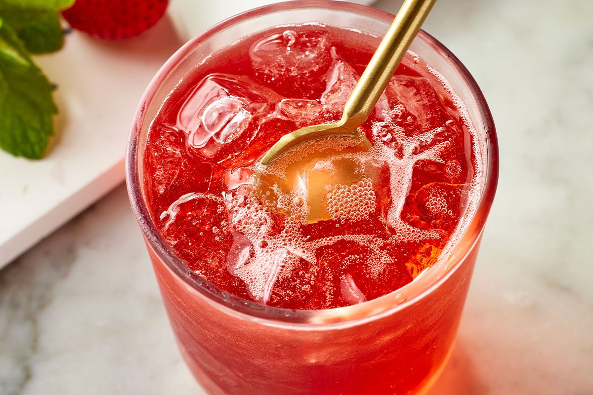 Overhead horizontal image of a glass of red beverage filled with ice and being stirred with a gold spoon. The drink appears to be bubbly. The image also shows a strawberry and mint leaf. | [Original] Stir to combine, garnish with mint leaves and fresh strawberries.