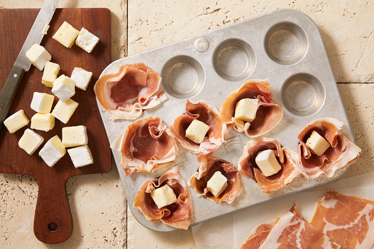 A muffin tin lined with slices of prosciutto, each holding a cube of brie cheese. Next to it, a wooden board with more brie cubes and a knife, and a few slices of prosciutto nearby.