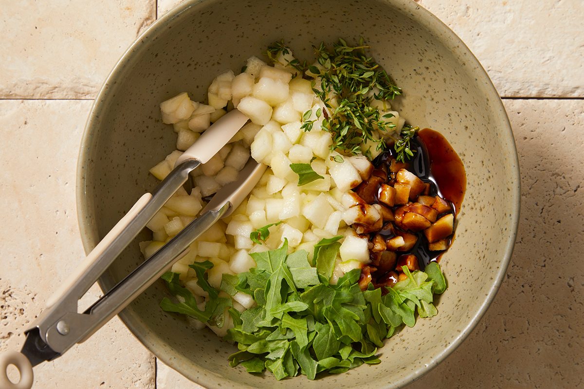 A bowl containing chopped onions, fresh green herbs, a leafy green, and a dark sauce, with a pair of tongs resting inside, on a tiled surface.