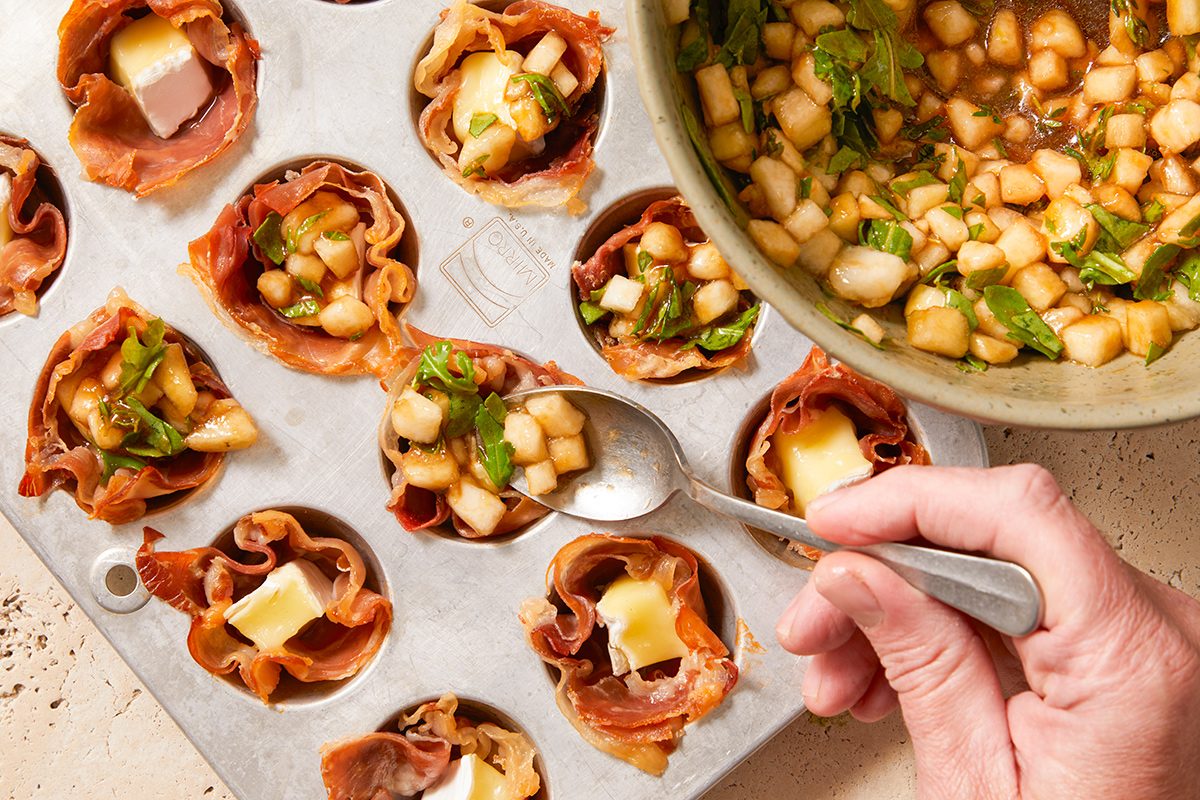 A hand spooning a diced pear and herb mixture into prosciutto-lined muffin tin cups filled with cheese, preparing savory appetizers.