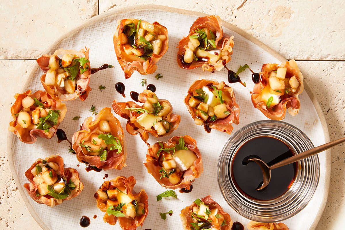 A glass plate with several small phyllo pastry cups filled with diced fruit, herbs, and cheese, served with a small bowl of dark dipping sauce and a spoon.