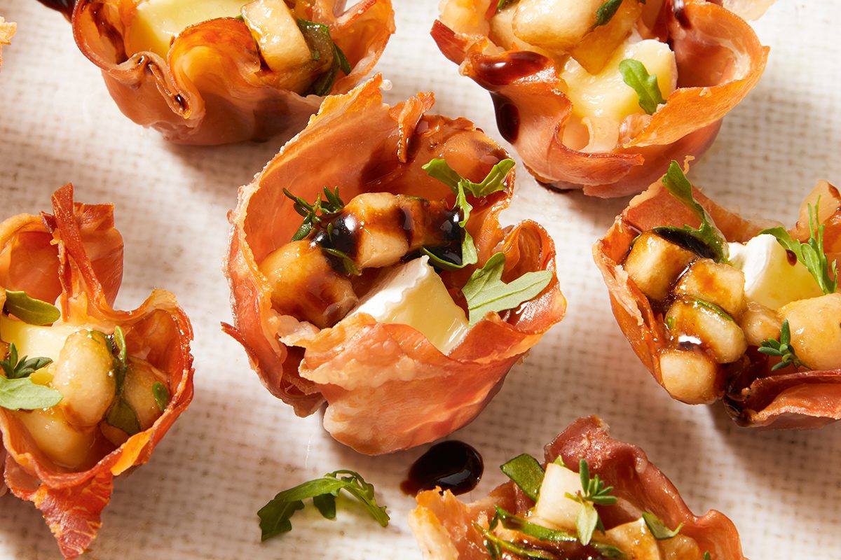 Mini prosciutto cups filled with cheese, diced pear, fresh herbs, and a drizzle of balsamic glaze, arranged on a light-colored textured surface.